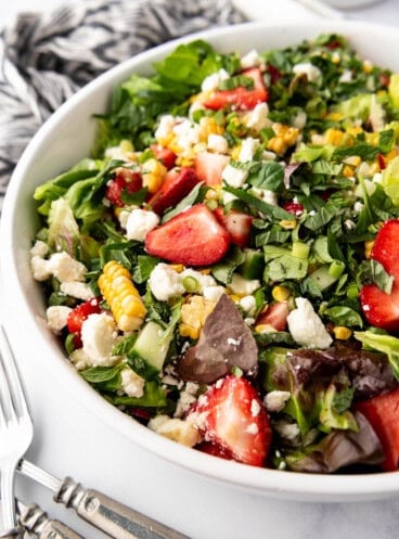 An image of a summer salad made with strawberries, corn, feta, and fresh herbs in a large white salad bowl.