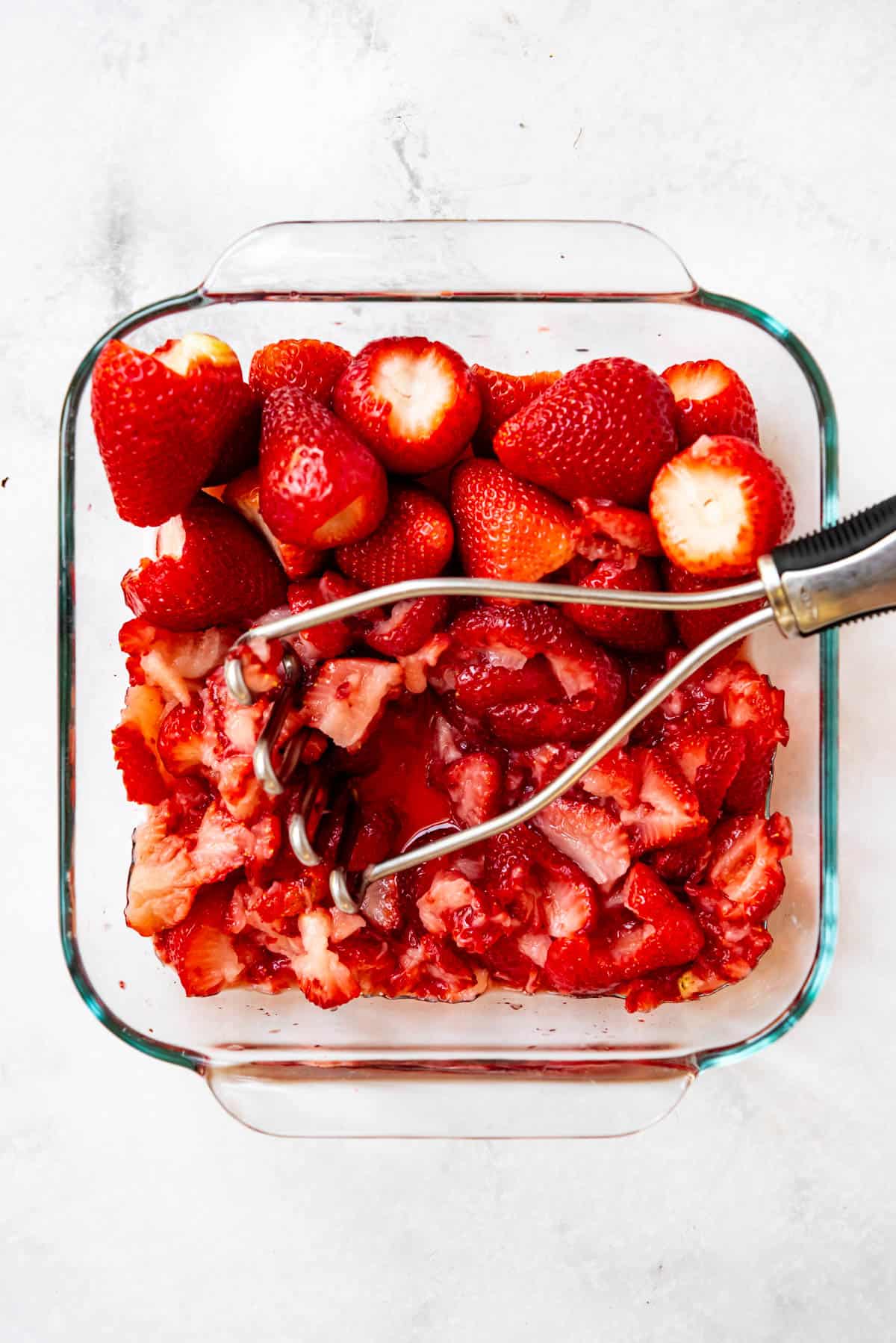 Strawberries in a glass baking dish with a potato masher.