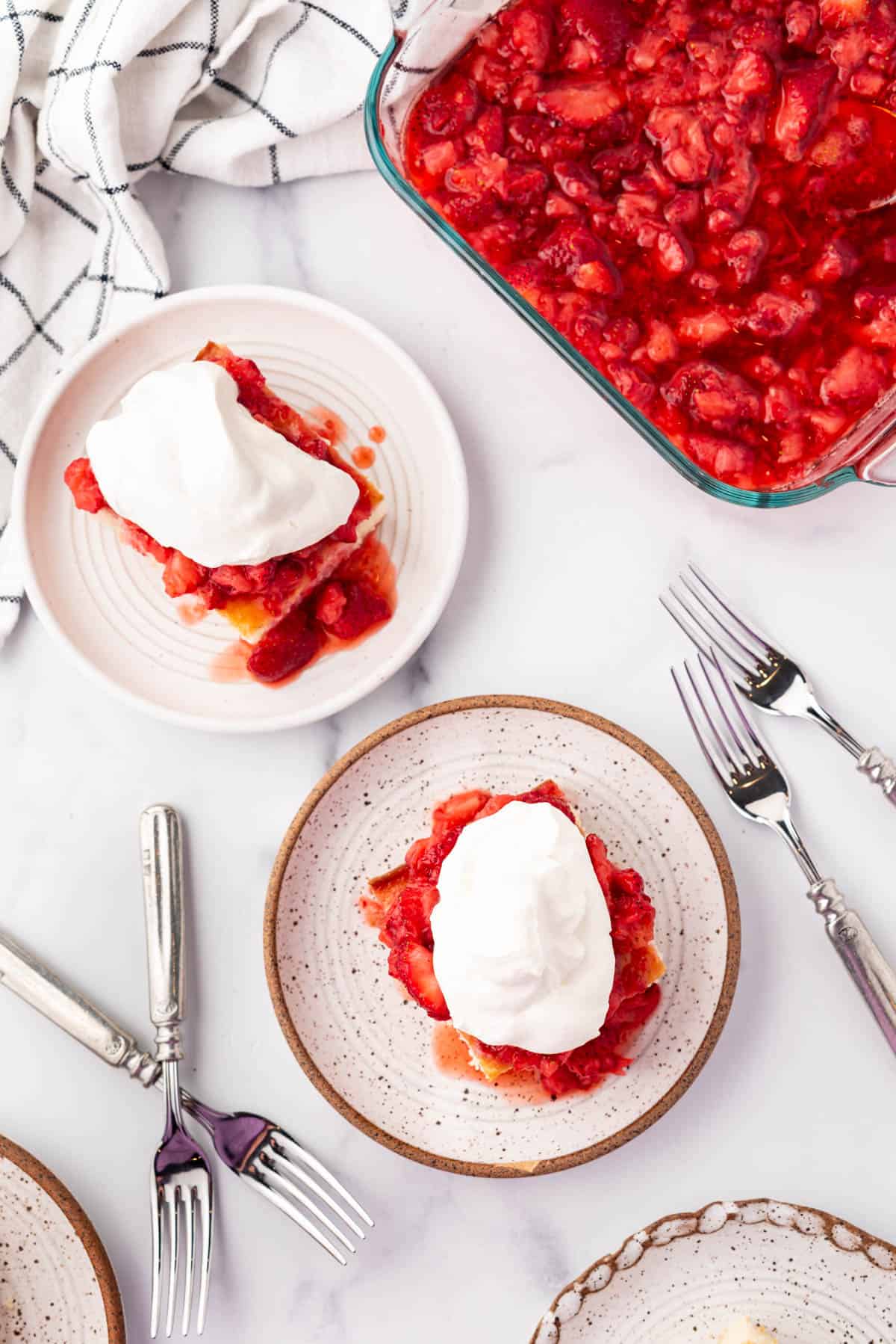 An overhead image of two slices of strawberry shortcake on plates next to forks and mashed strawberries.