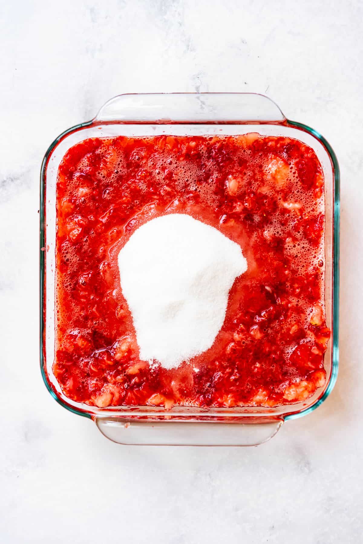 Adding granulated sugar to mashed strawberries in a glass baking dish.