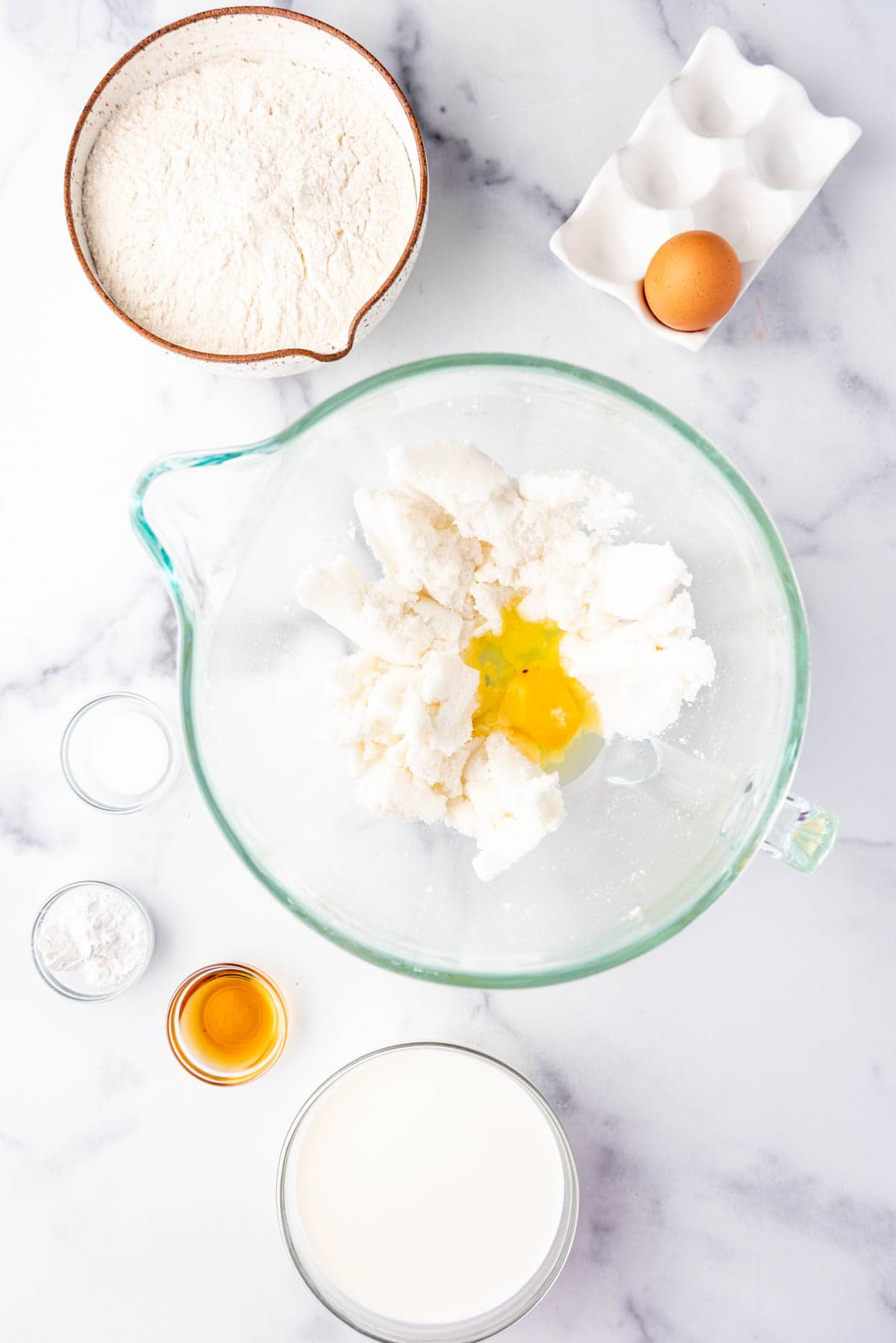 Adding an egg to creamed shortening and sugar in a large glass mixing bowl with more ingredients surrounding the bowl.