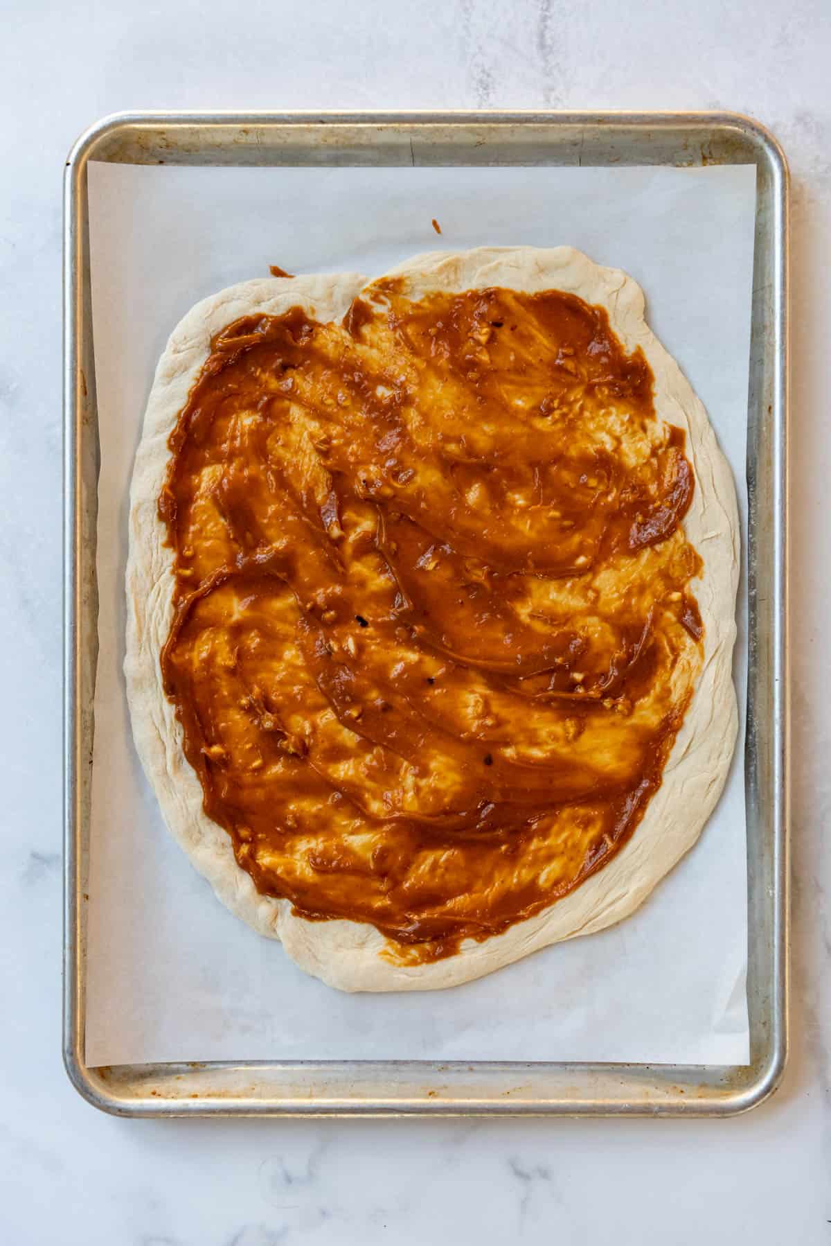 Spreading peanut sauce on a pizza crust on a baking sheet lined with parchment paper.