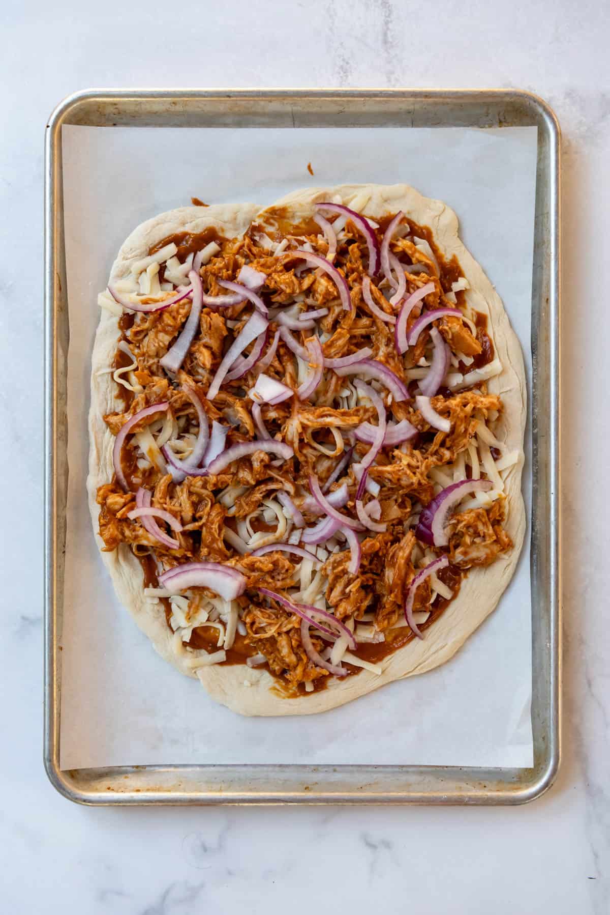 An unbaked Thai chicken pizza topped with shredded chicken and sliced red onion.