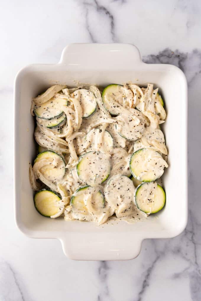 Layering sliced zucchini and onions in a creamy sauce in a square white baking dish.