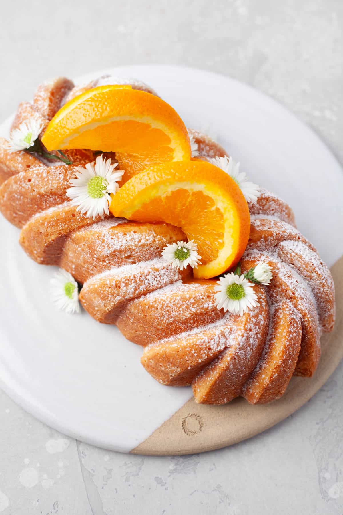 Overhead view of Orange Loaf Cake on a white serving plate with slices of orange and small white flowers garnishing it.