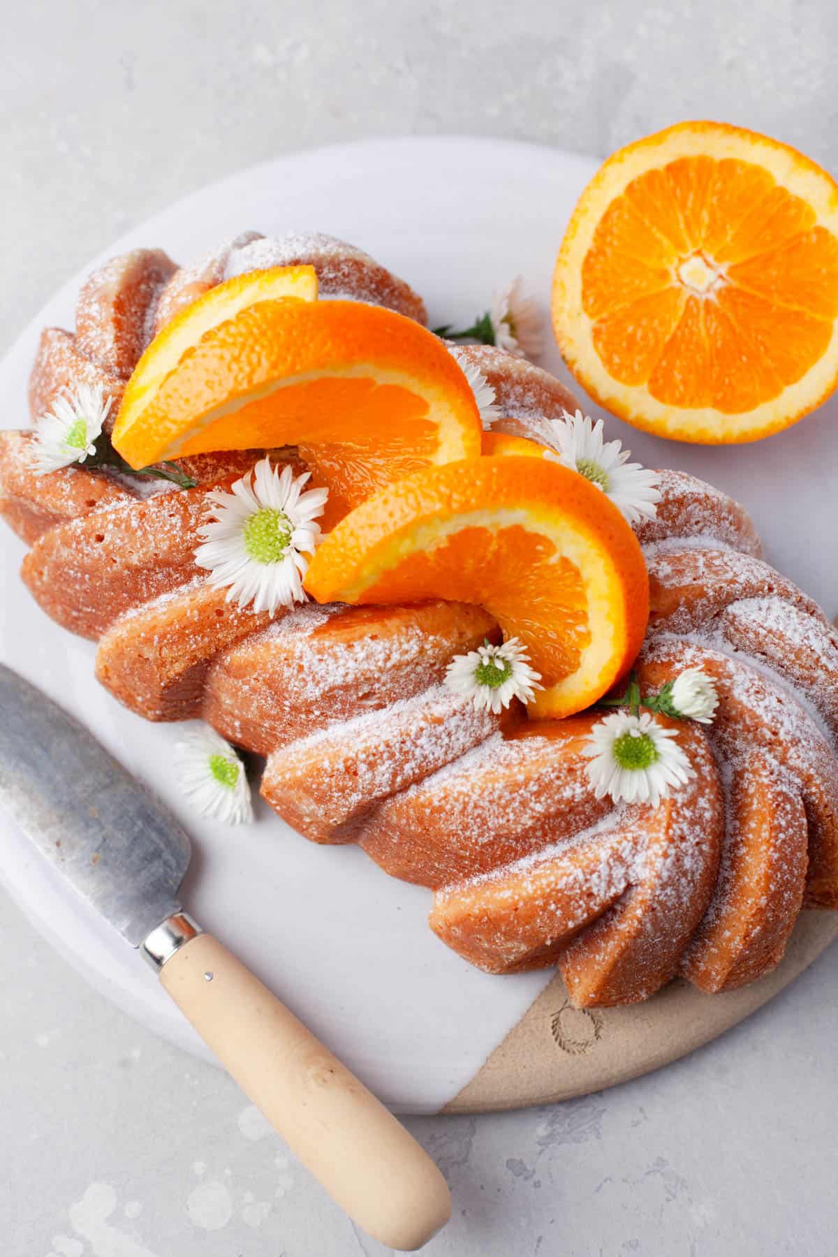 Overhead view of Orange Loaf Cake on a white serving plate with slices of orange and small white flowers garnishing it.