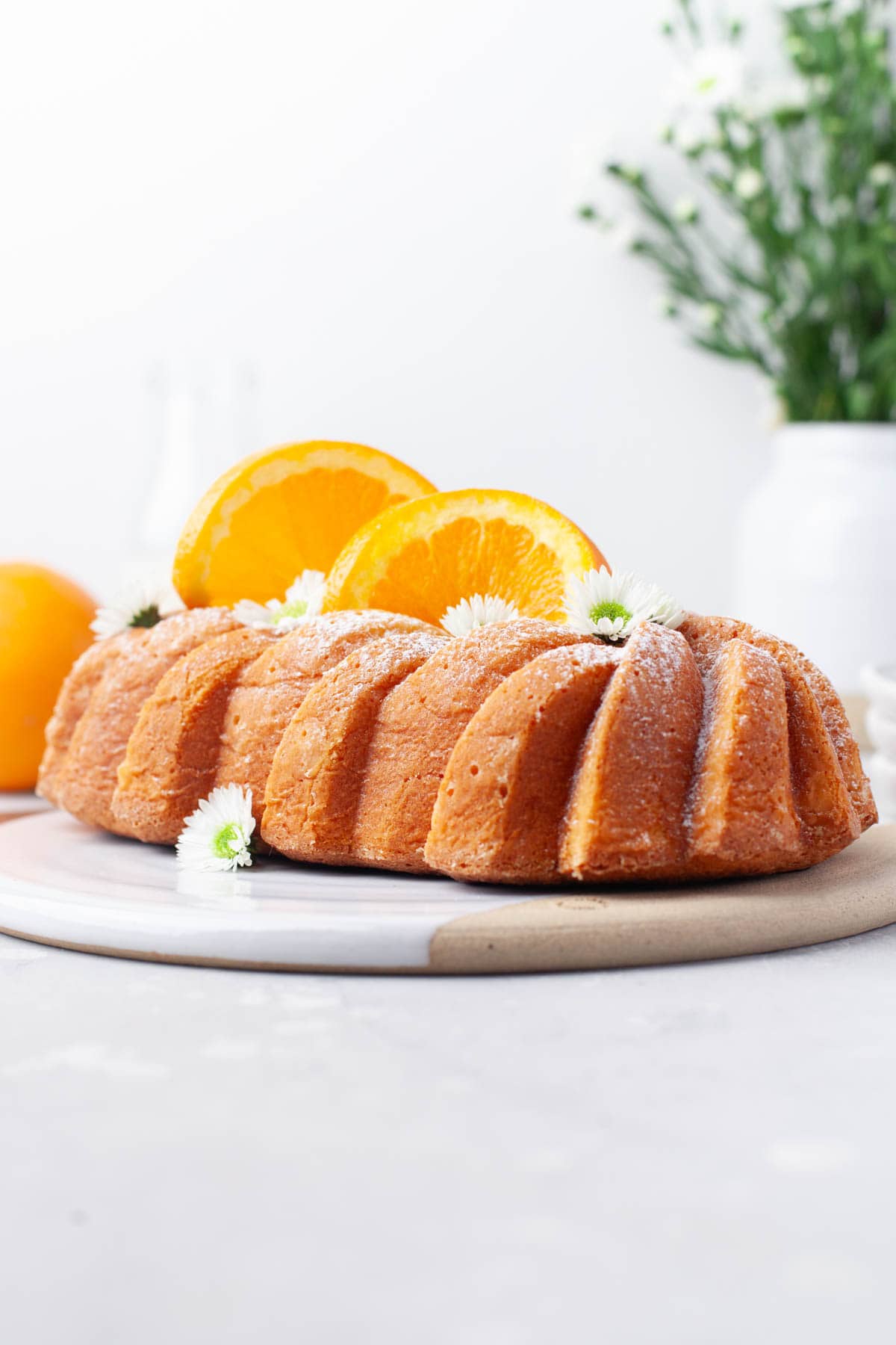 Side view of Orange Loaf Cake on a white serving plate with slices of orange and small white flowers garnishing it.