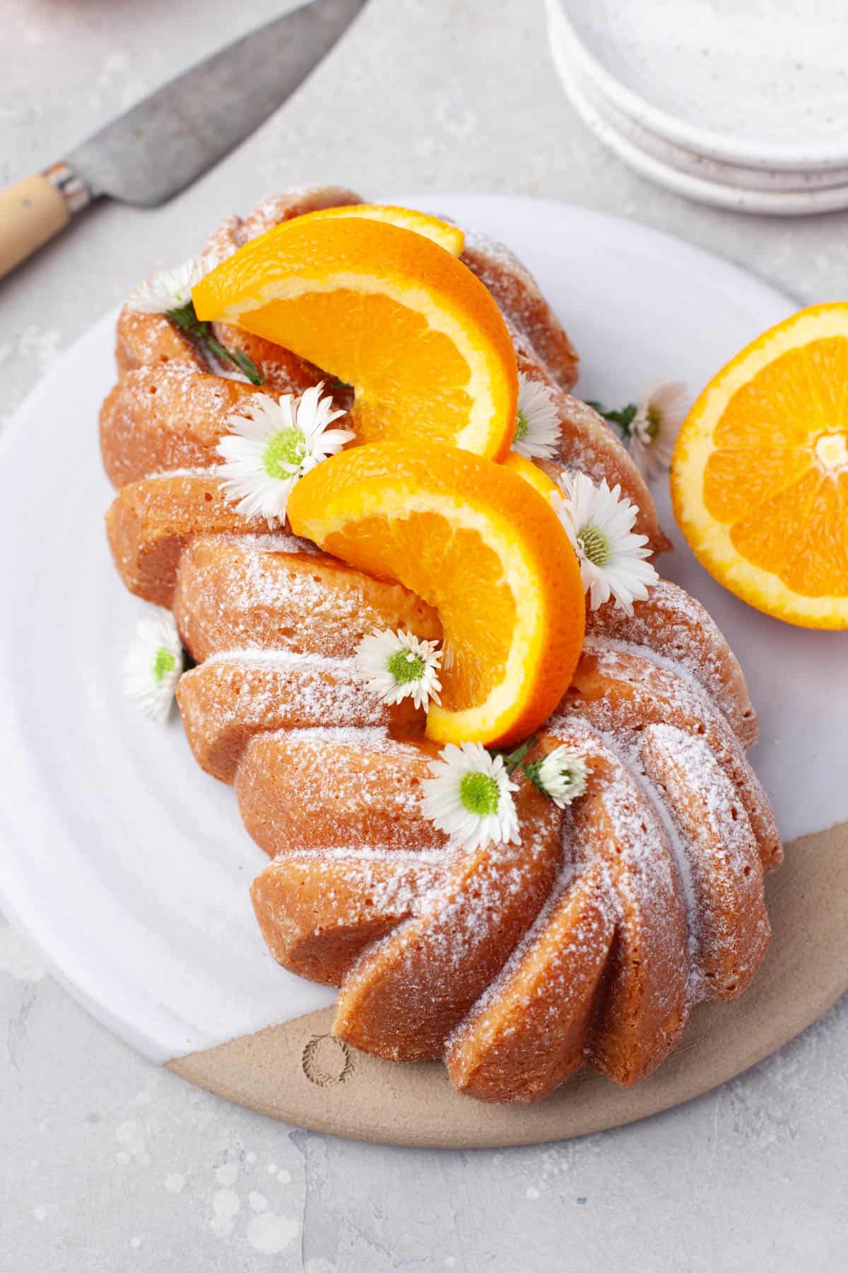 Overhead view of Orange Loaf Cake on a white serving plate with slices of orange and small white flowers garnishing it.