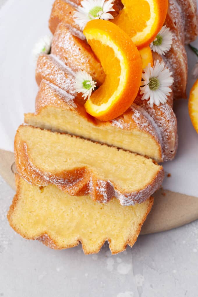 Overhead view of Orange Loaf Cake on a white serving plate with slices of orange and small white flowers garnishing it.