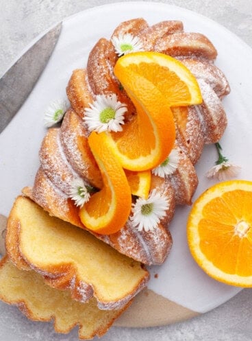 Overhead view of Orange Loaf Cake on a white serving plate with slices of orange and small white flowers garnishing it.