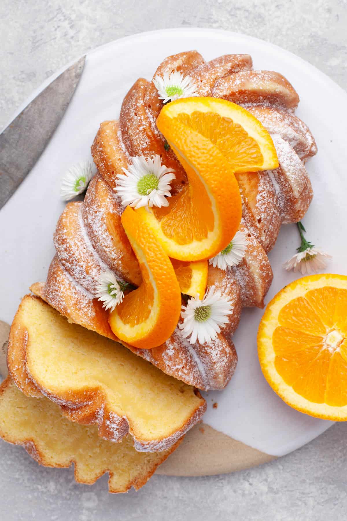 Vue de dessus d'un gâteau de pain à l'orange sur une assiette blanche avec des tranches d'orange et de petites fleurs blanches en garniture.