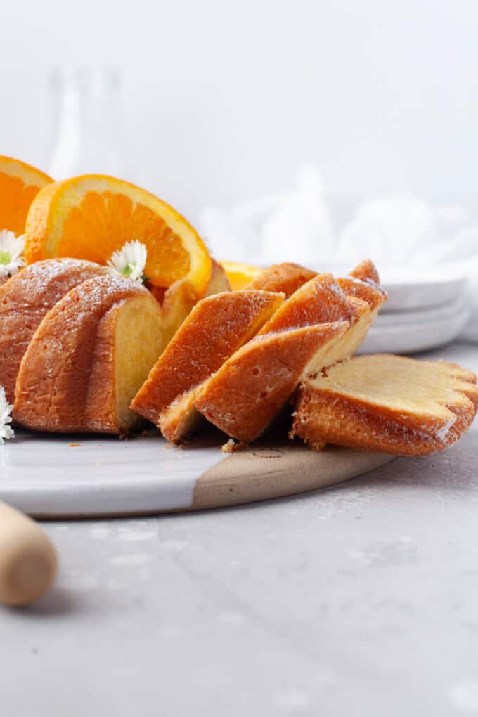 Vue de côté d'un gâteau de pain à l'orange sur une assiette blanche avec des tranches d'orange et de petites fleurs blanches en garniture.