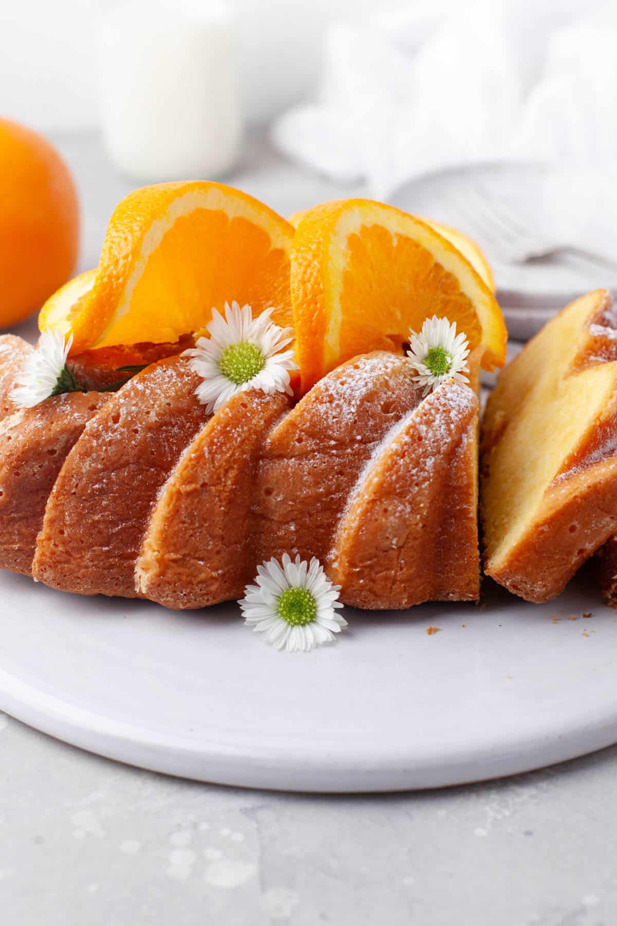 Side view of Orange Loaf Cake on a white serving plate with slices of orange and small white flowers garnishing it.