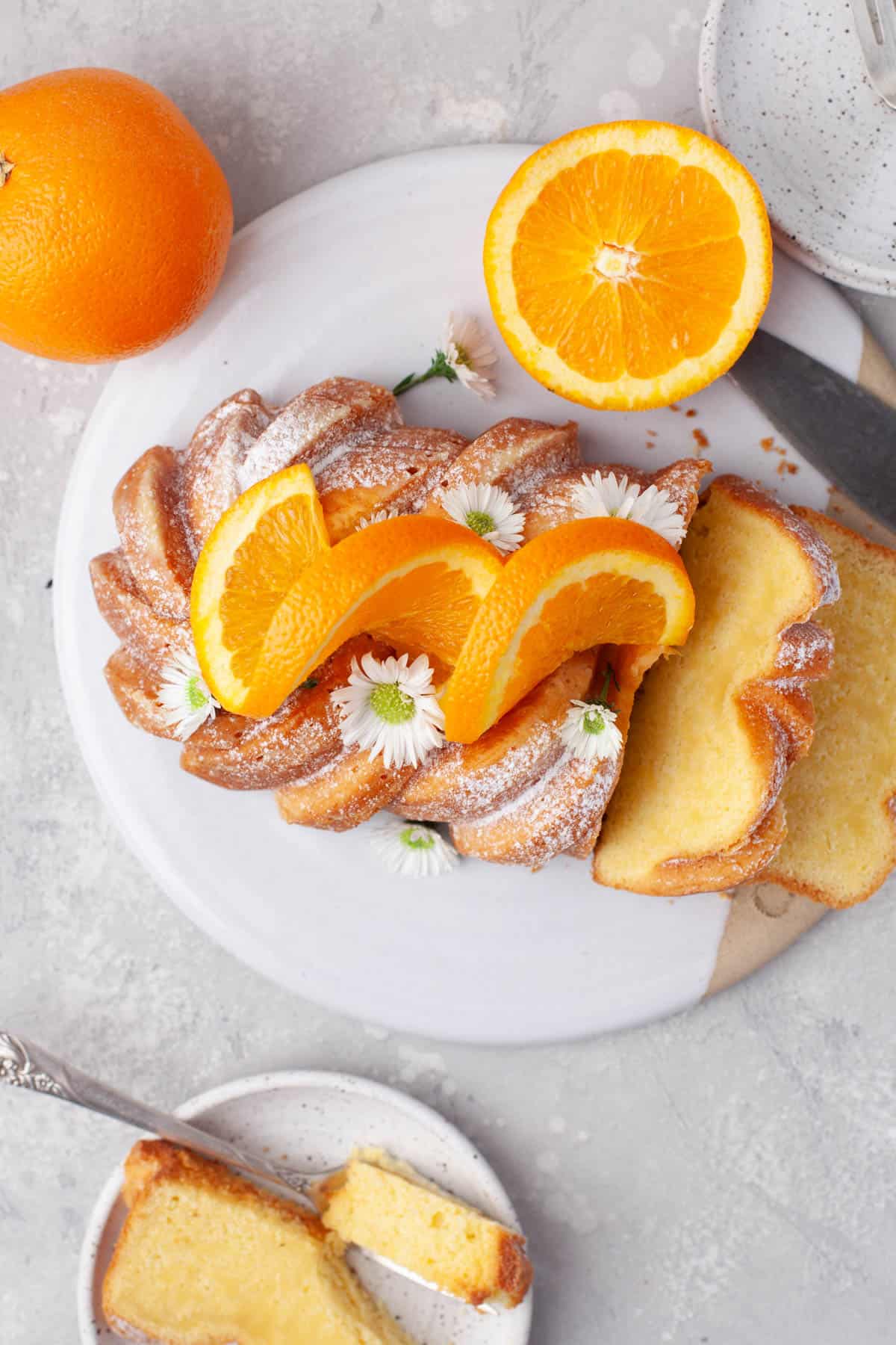 Overhead view of Orange Loaf Cake on a white serving plate with slices of orange and small white flowers garnishing it.