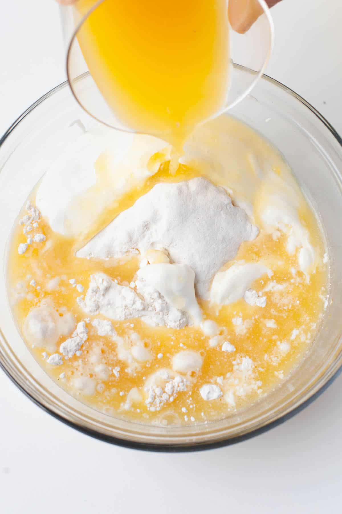 Overhead view of orange juice being added to a glass mixing bowl with sour cream, flour and baking powder in it.