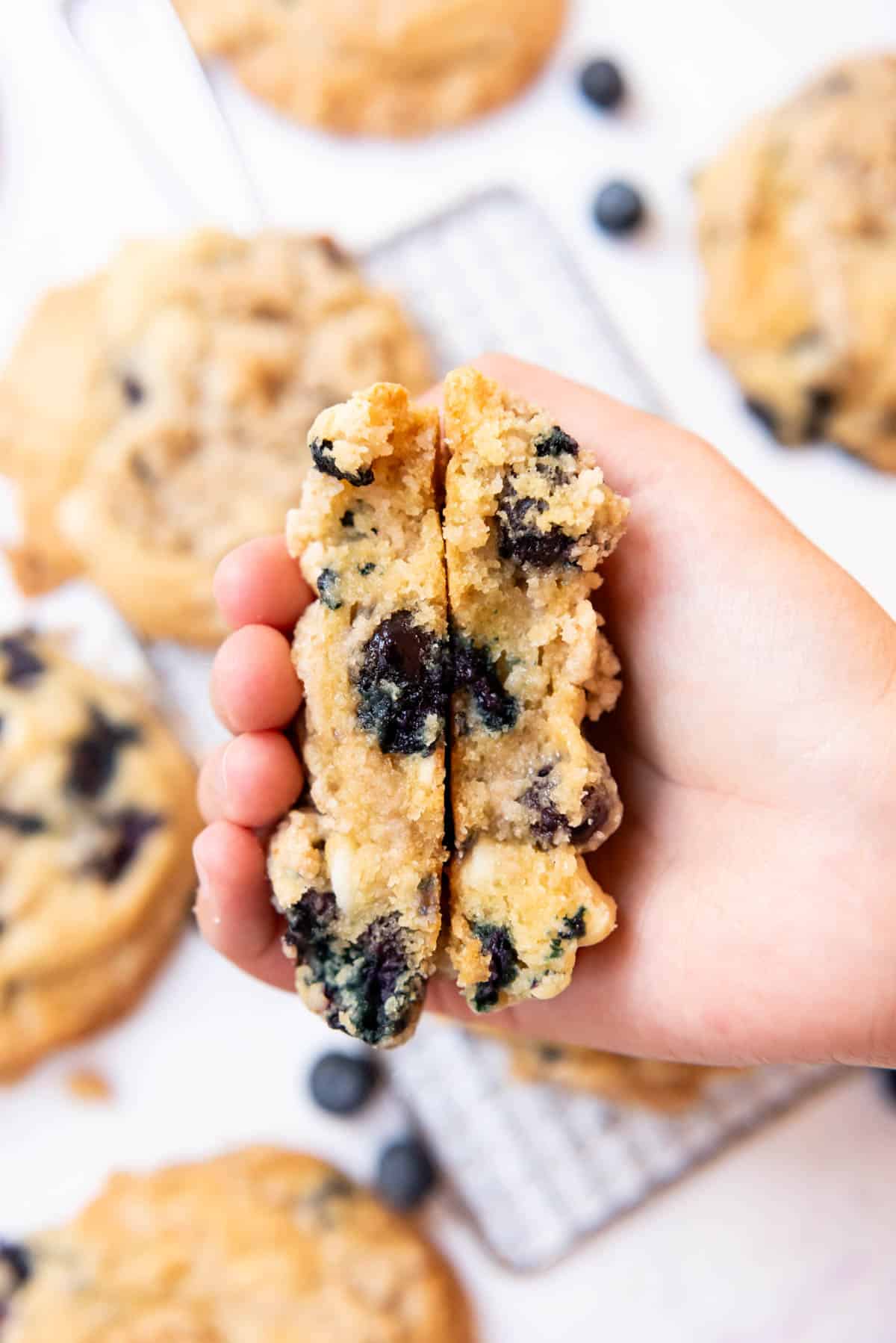 An overhead image of a hand holding two halves of a blueberry muffin cookie.