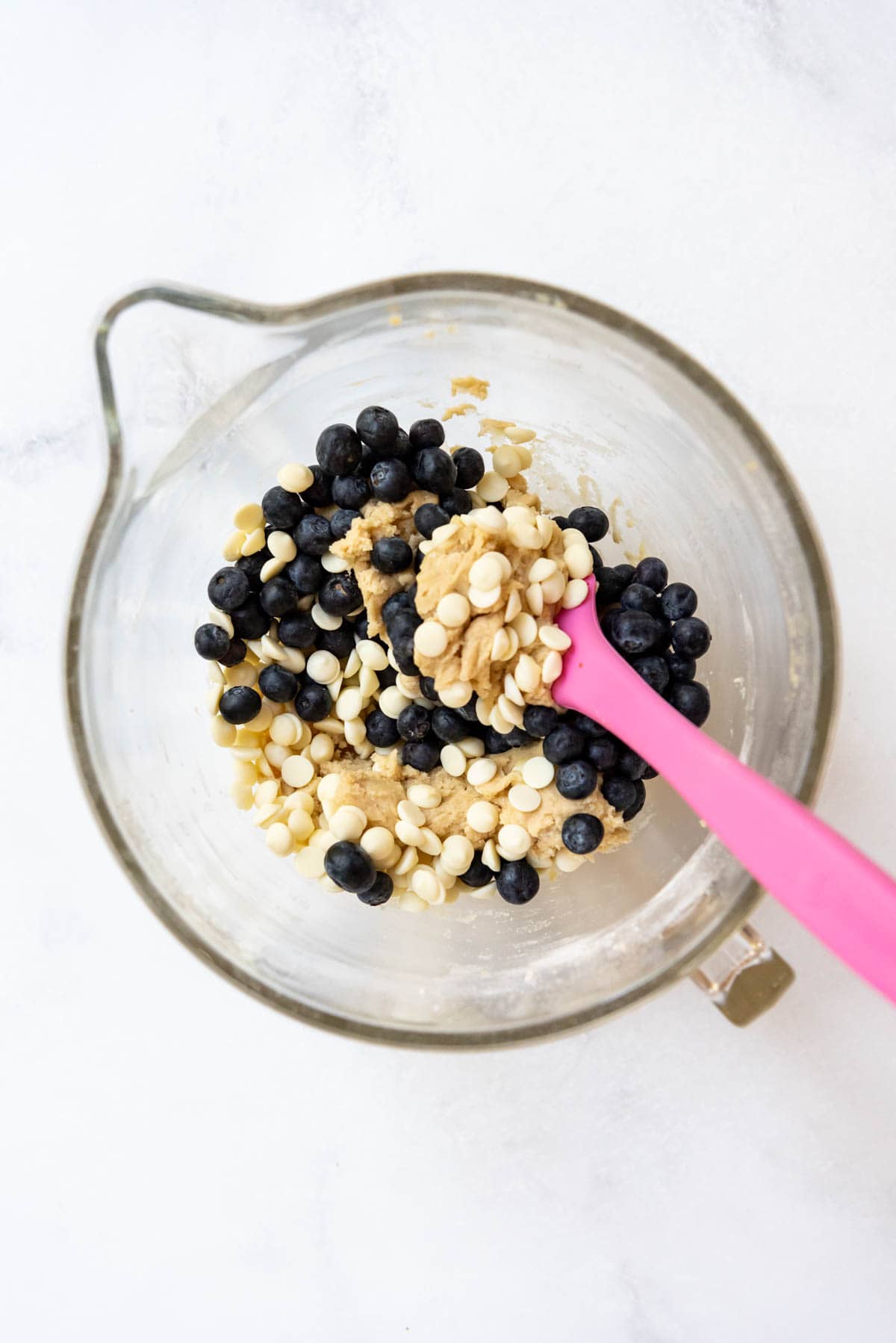 Folding white chocolate chips and fresh blueberries into cookie dough in a glass mixing bowl.