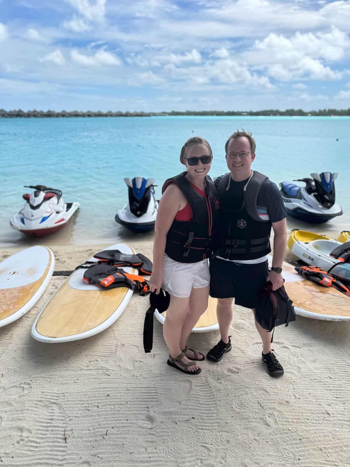 A couple in life jackets in front of paddle boards and jet skis.