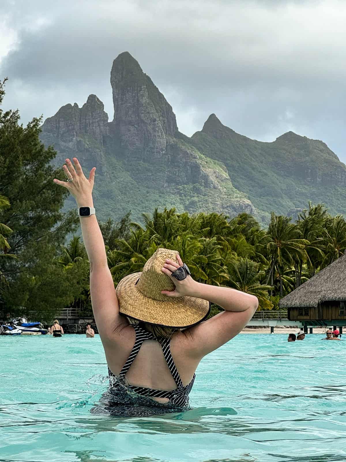 An image of a woman holding on to her hat in the ocean while facing the island of Bora Bora.