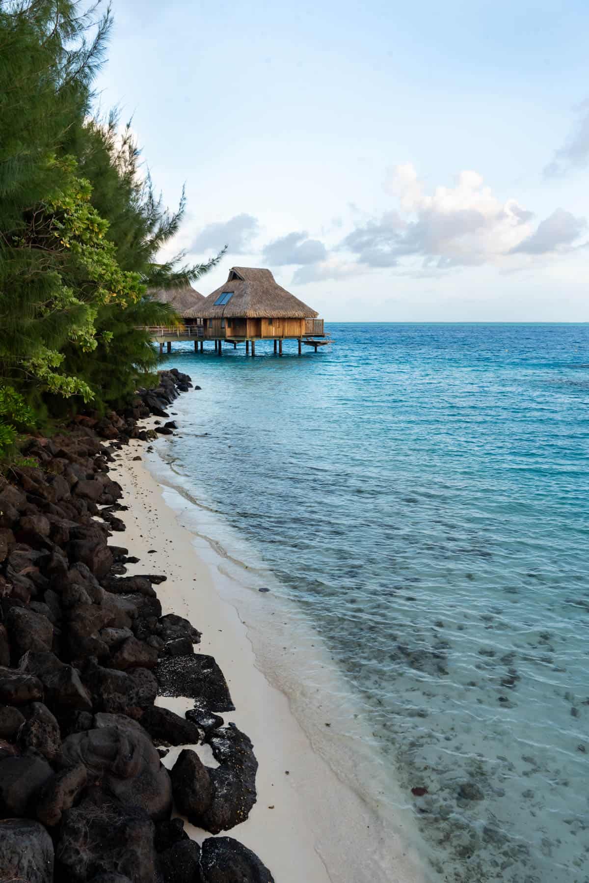 A narrow white sandy beach lined with black volcanic rock beside a bright blue ocean with a bungalow in the background.