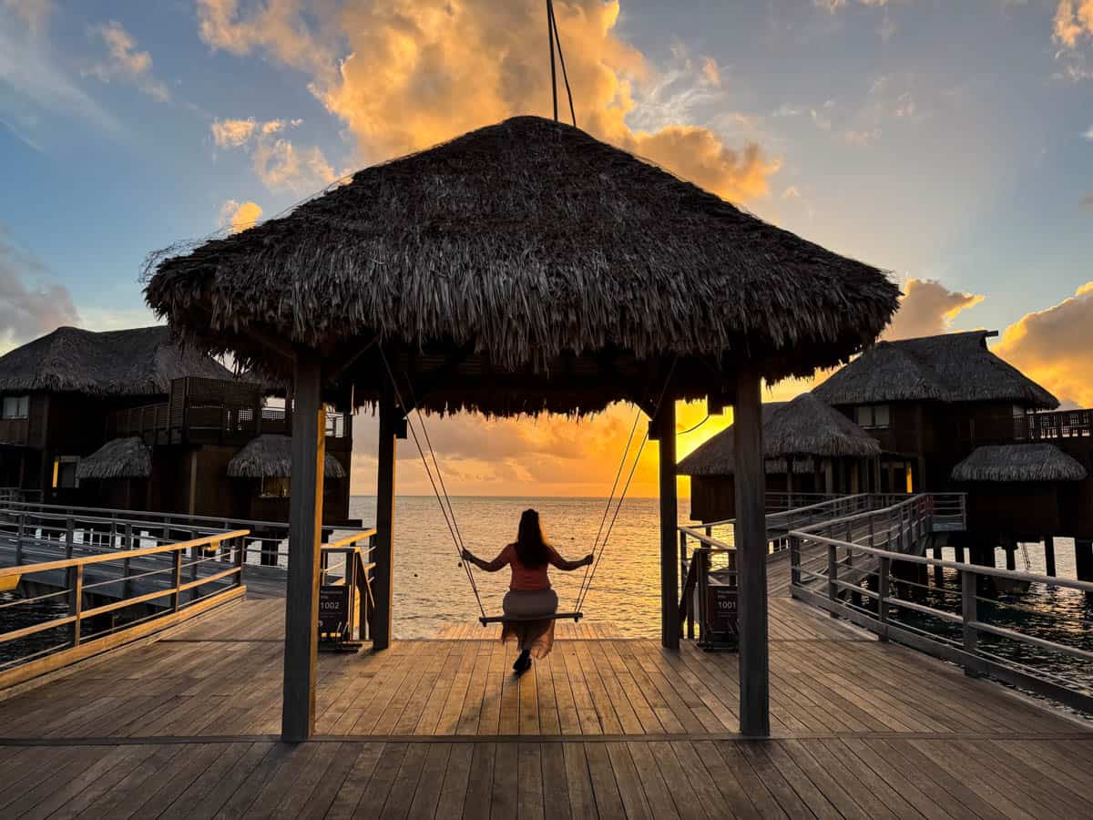 An image of a woman on a hanging bench looking at the sunset in Tahiti.