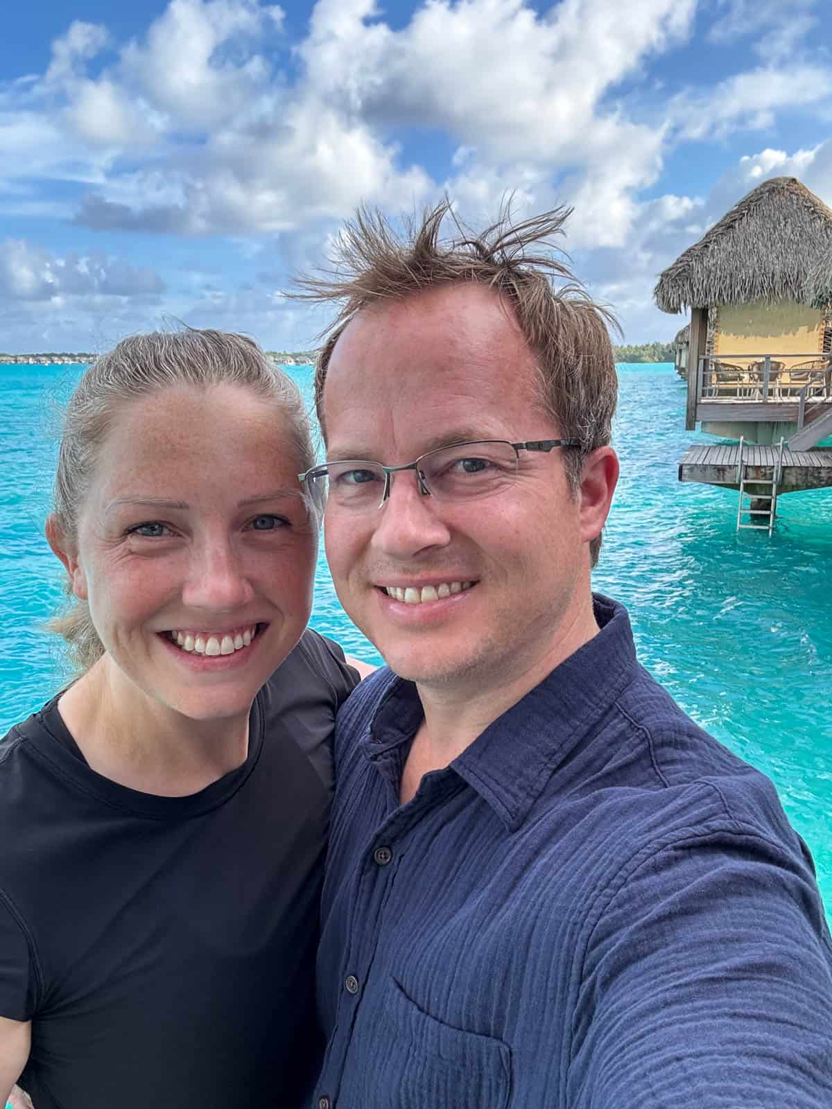 An image of a couple taking a selfie on a private balcony in Bora Bora with an over-the-water bungalow behind them.