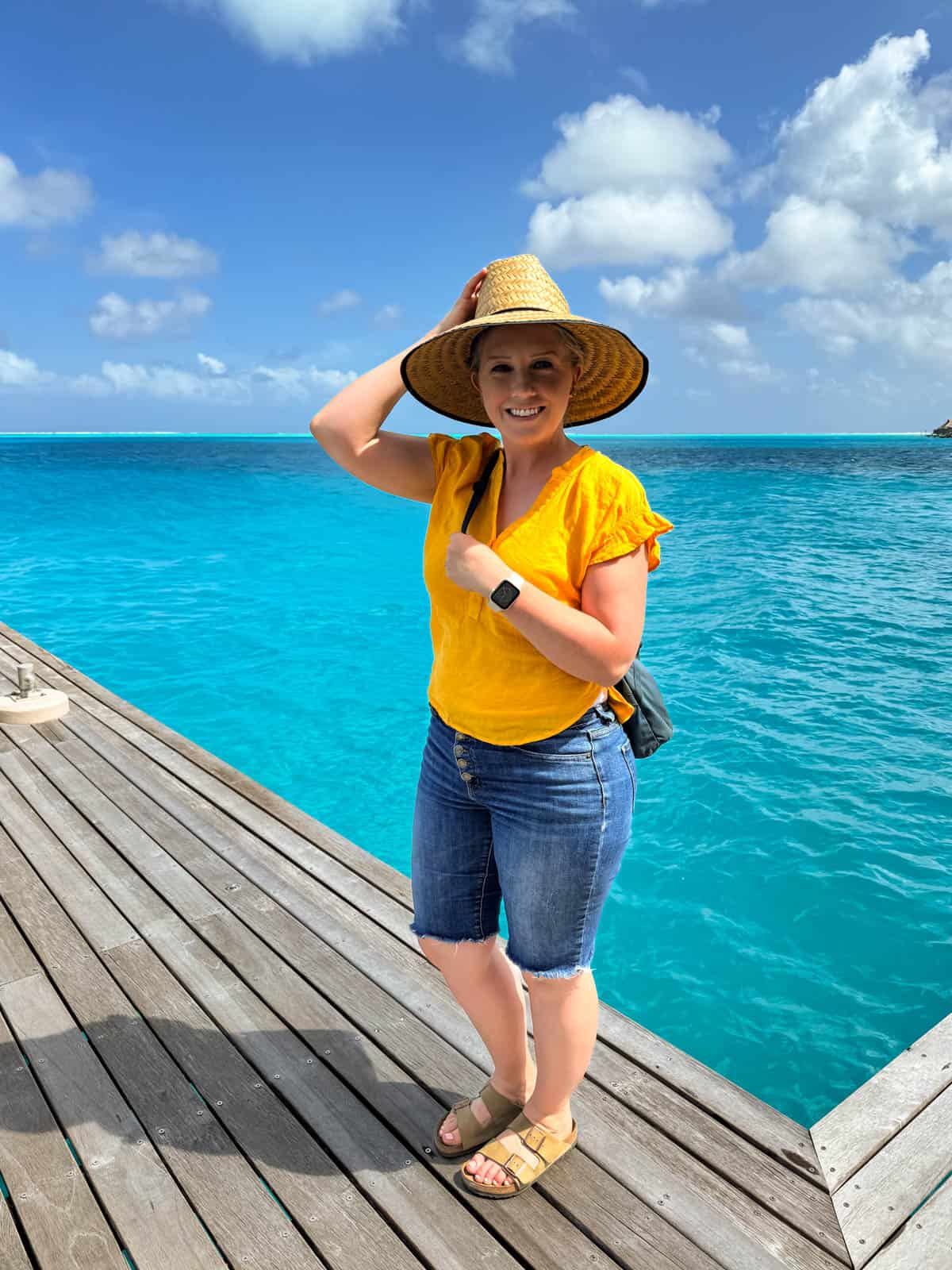 An image of a woman in a yellow shirt and sun hat on a dock in front of turquoise blue water in French Polynesia.