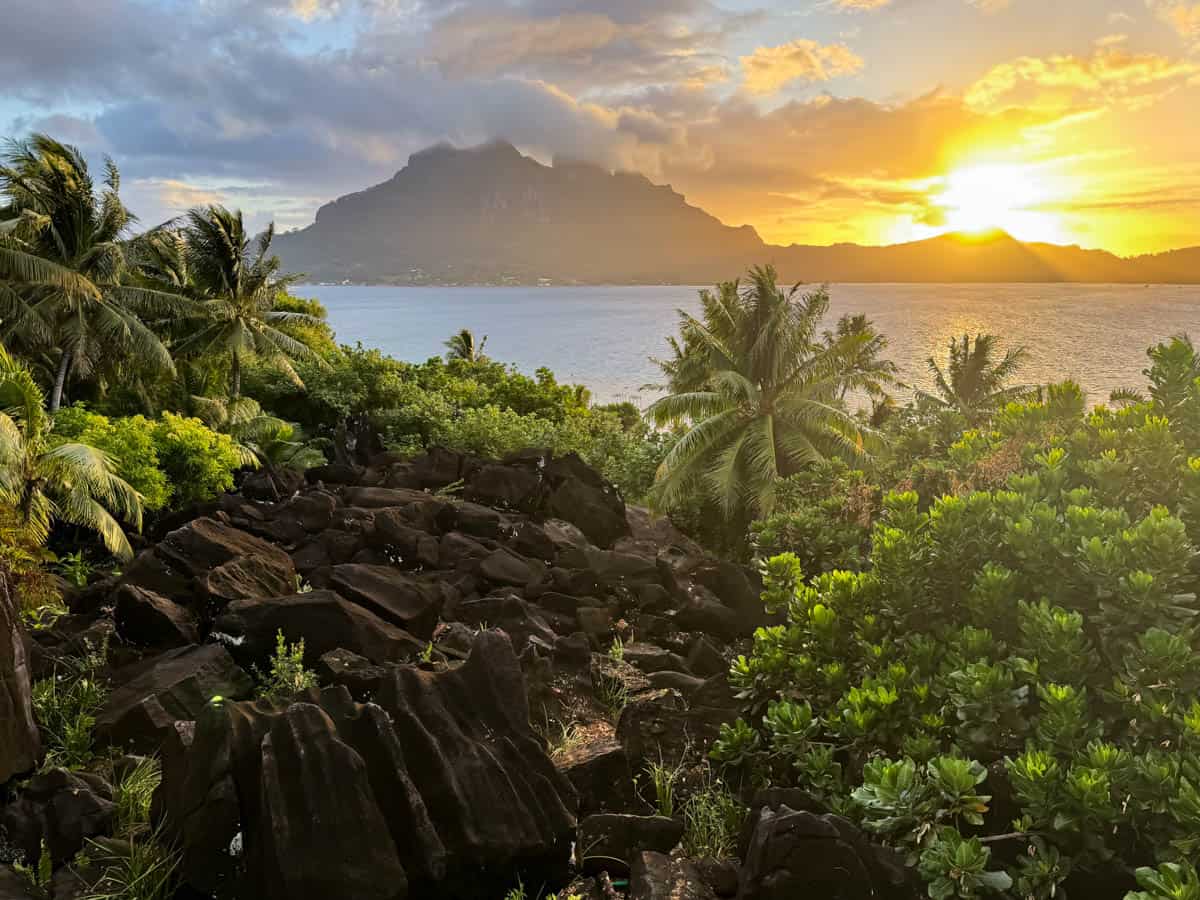 An image of the sun setting over the island of Bora Bora in French Polynesia with lava rocks and palm trees in the foreground.