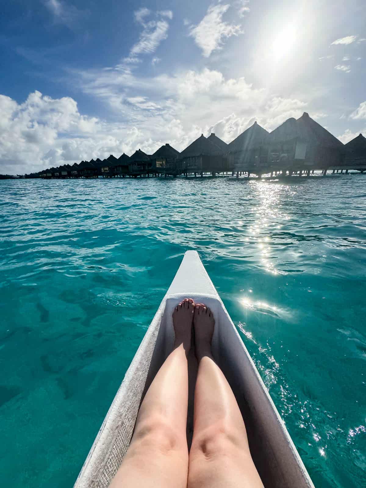 An image of a person's legs and feet in a white canoe on turqouise water heading to huts at the St. Regis Bora Bora resort.