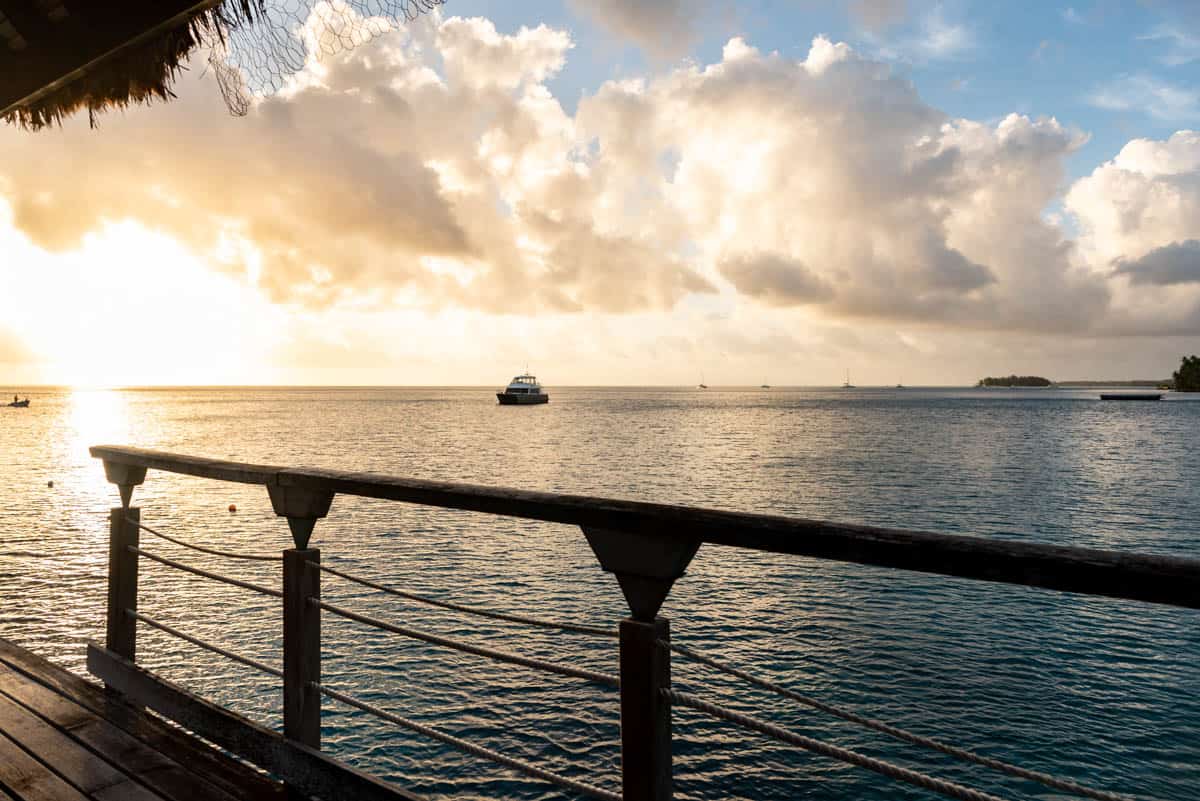 Boats on the ocean at sunset with a deck railing in the foreground.