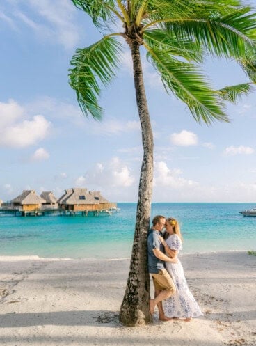 An image of a couple on a beach leaning against a palm tree.