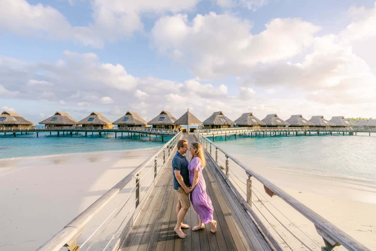 An image of a couple holding hands on a wooden walkway with over-the-water bungalows in the background.