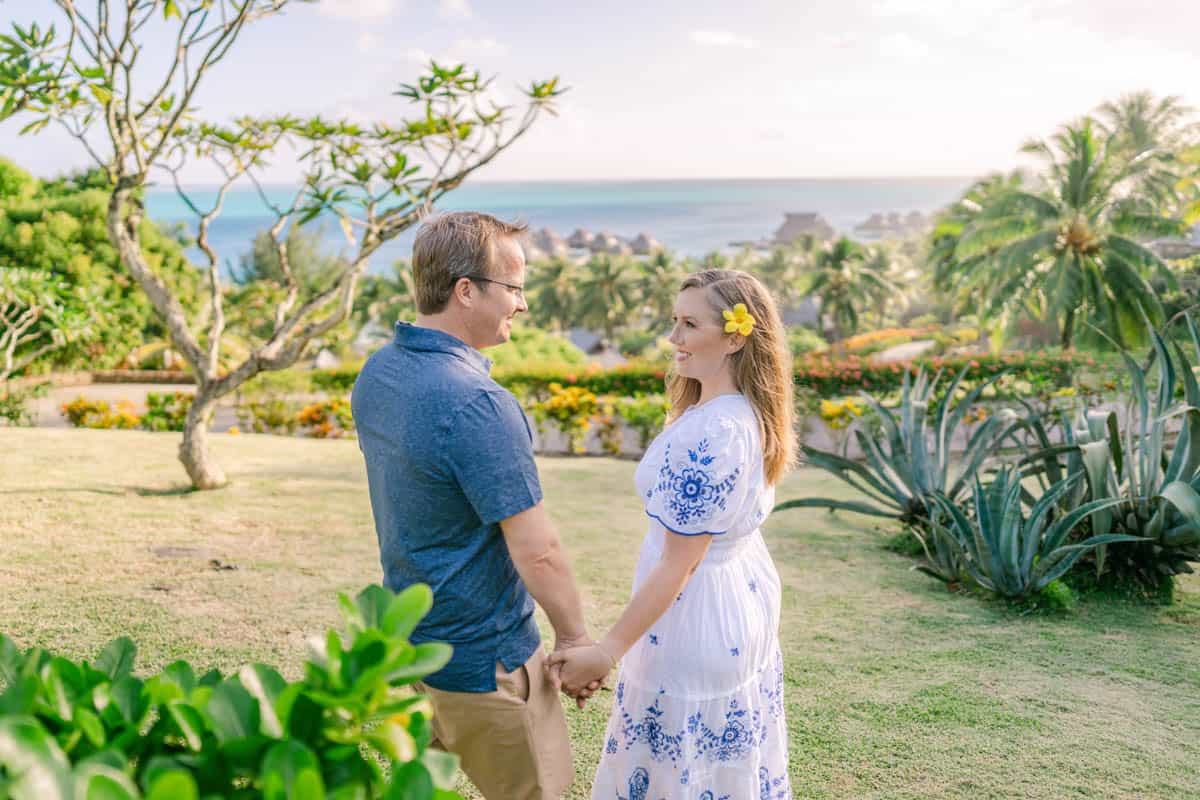 An image of a woman with a yellow flower in her hair and white dress holding hands with a man in a blue shirt in Bora Bora.