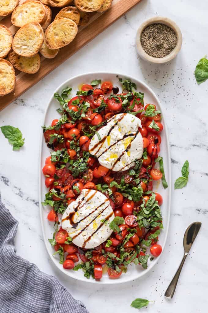 Burrata caprese drizzled with balsamic glaze next to sliced and toasted baguette rounds.