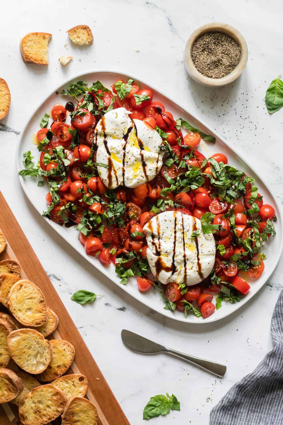 An overhead image of burrata caprese on a white serving platter with toasted bread nearby on a wooden cutting board.