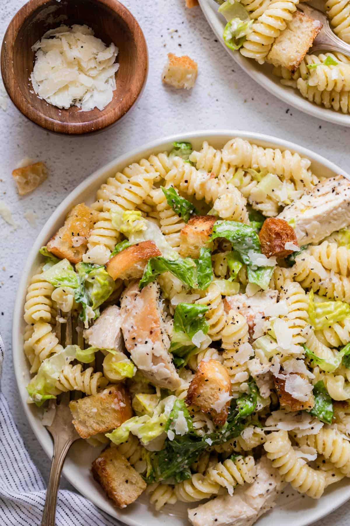 An overhead image with a plate of pasta salad with chicken in the bottom right hand corner with a small bowl of parmesan cheese above it.