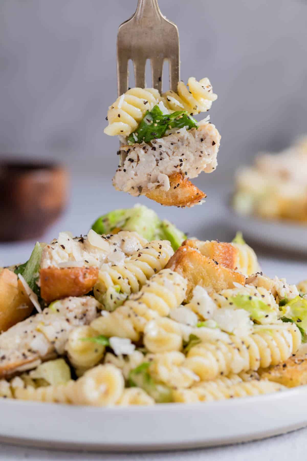 A fork lifting a bite of chicken caesar pasta salad from a plate.