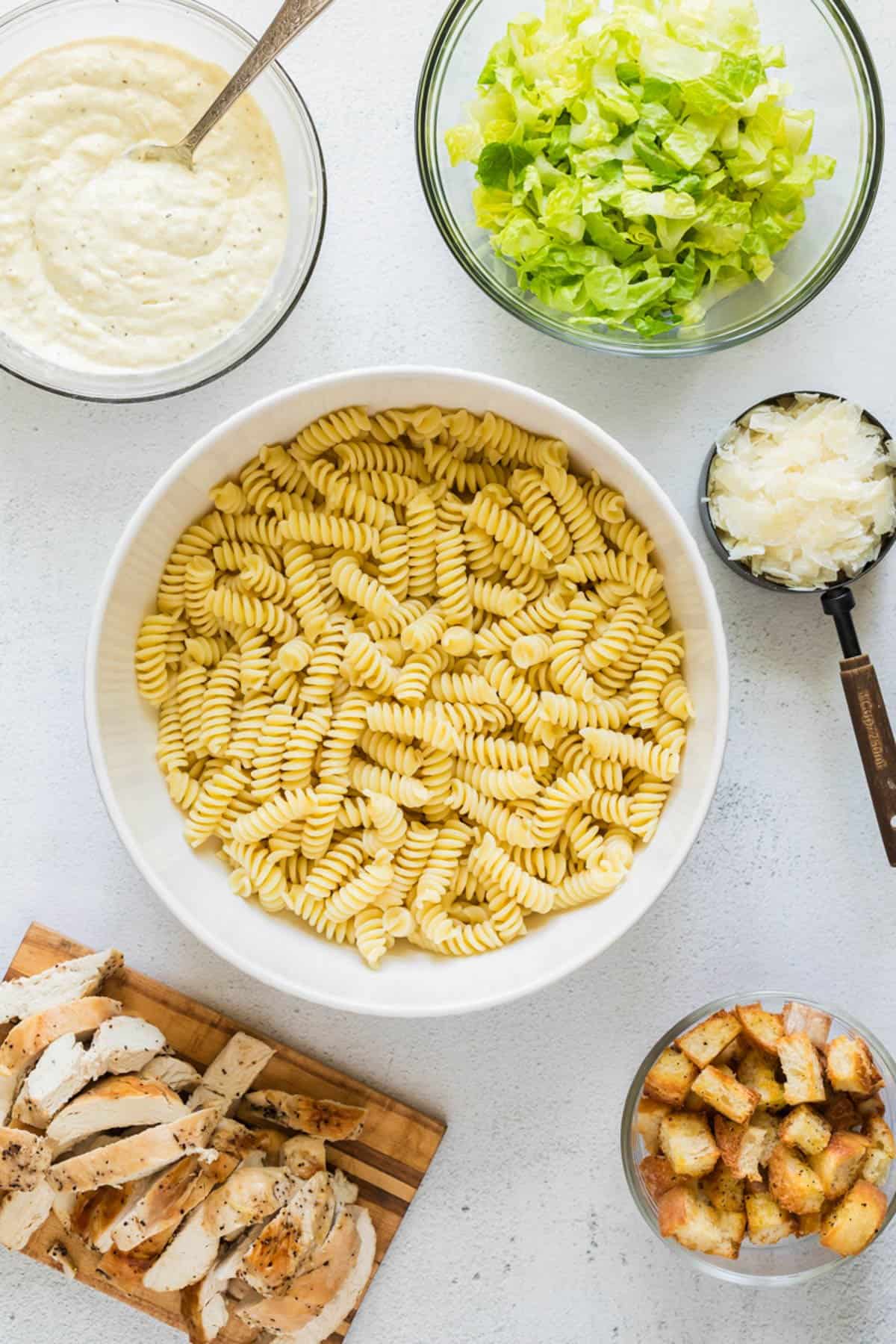 Cooked fusili pasta in a large serving bowl with ingredients nearby for making a chicken caesar pasta salad.