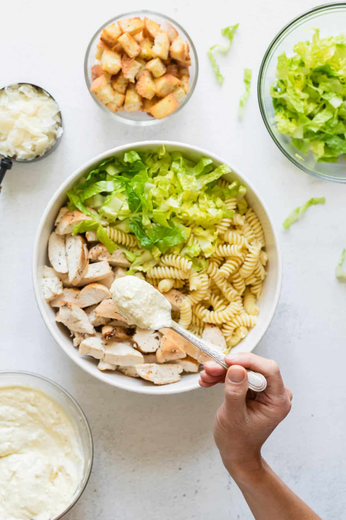 Adding salad dressing and other ingredients to make a pasta salad in a large bowl with croutons and chopped romaine lettuce nearby.