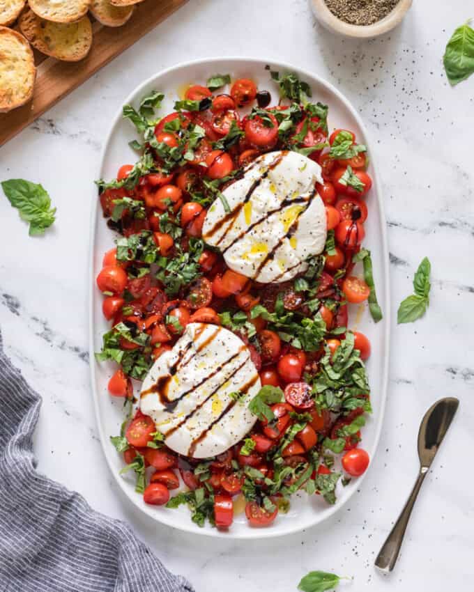 Burrata caprese drizzled with balsamic glaze next to sliced and toasted baguette rounds.