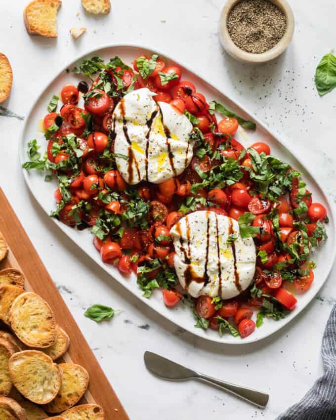 An overhead image of burrata caprese on a white serving platter with toasted bread nearby on a wooden cutting board.