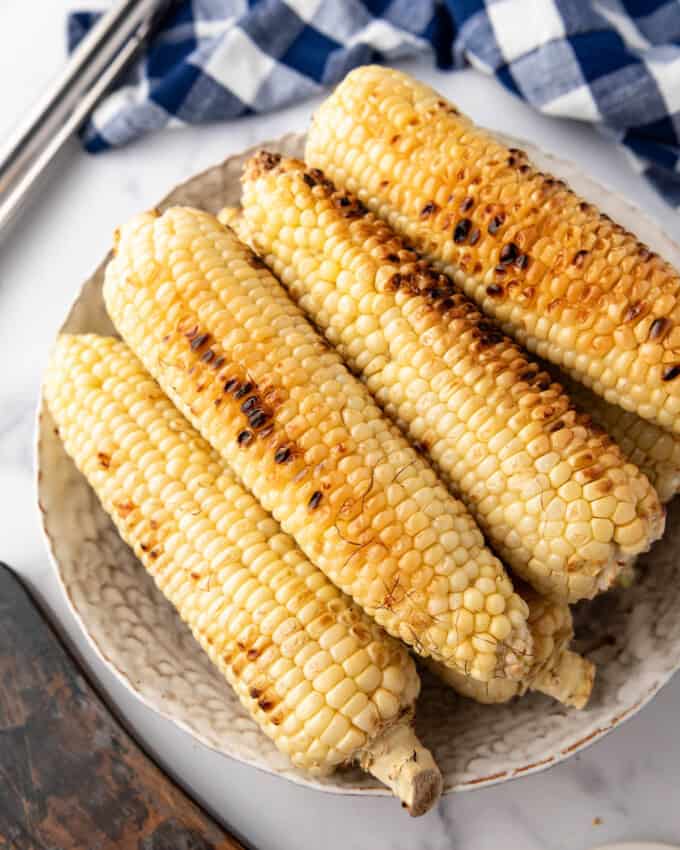 An overhead image of slightly charred ears of grilled corn on the cob with a blue and white checked cloth and tongs behind them.