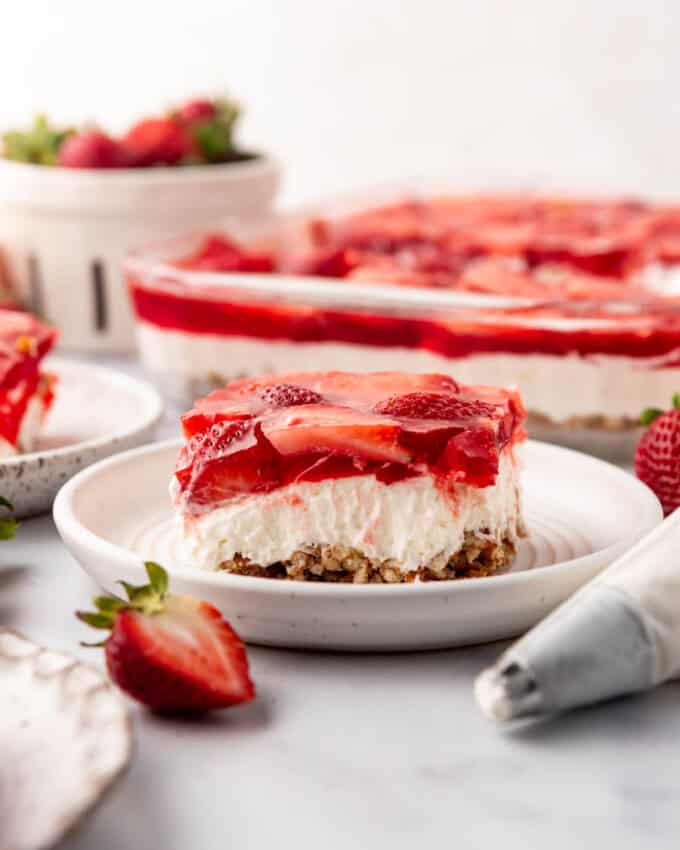 A slice of strawberry pretzel salad dessert on a plate in front of the pan of jello salad.