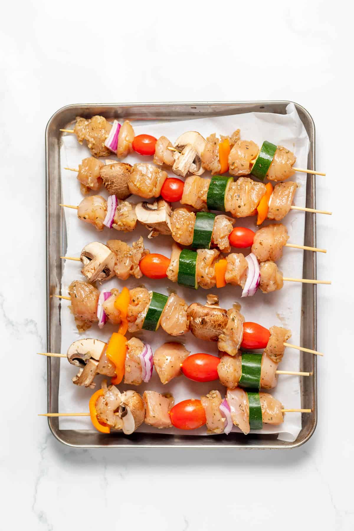 Wooden skewers holding marinated chicken and vegetables on a baking sheet.