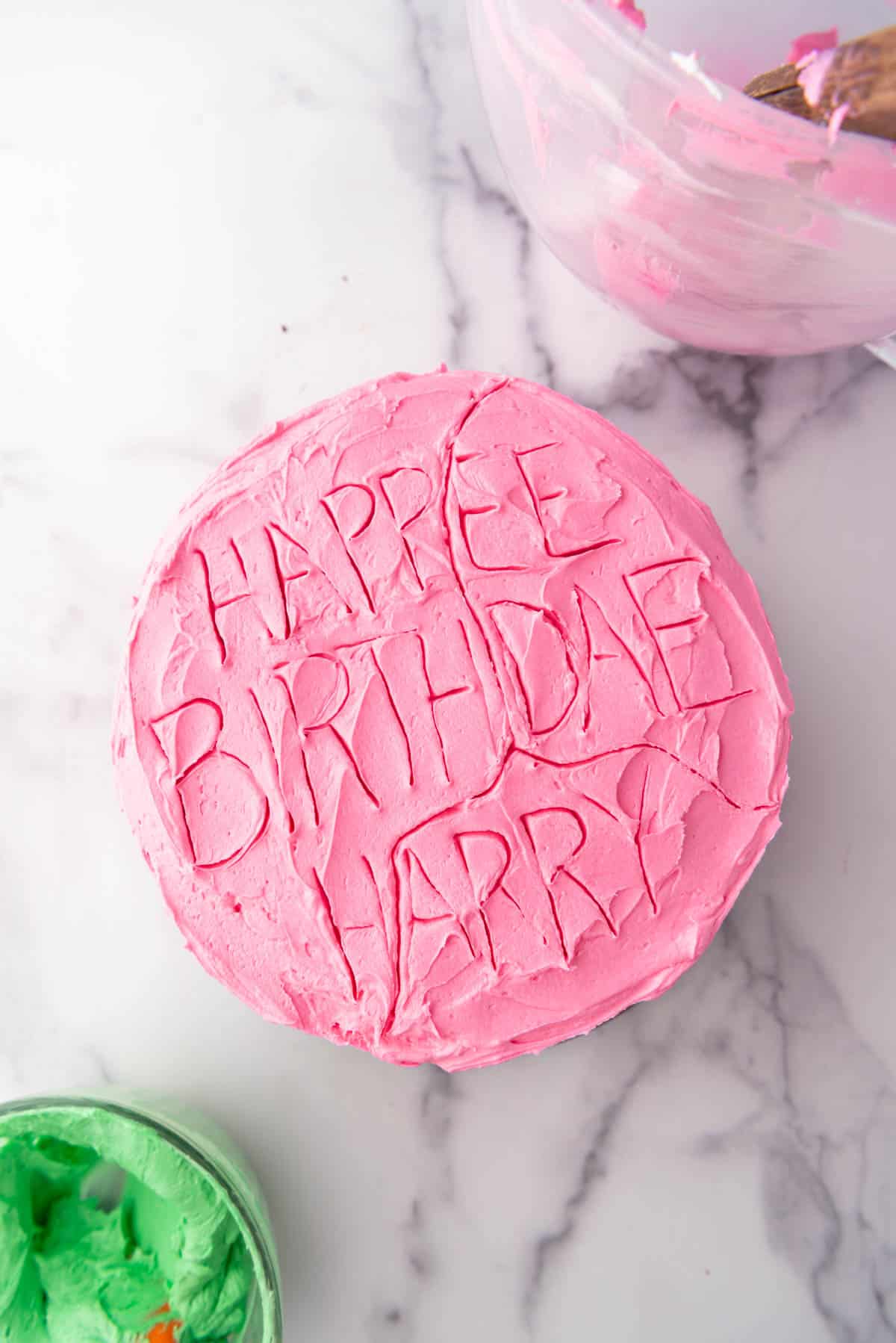 An image of a pink frosted cake with letters traced on top using a toothpick as a guide for piping frosting.