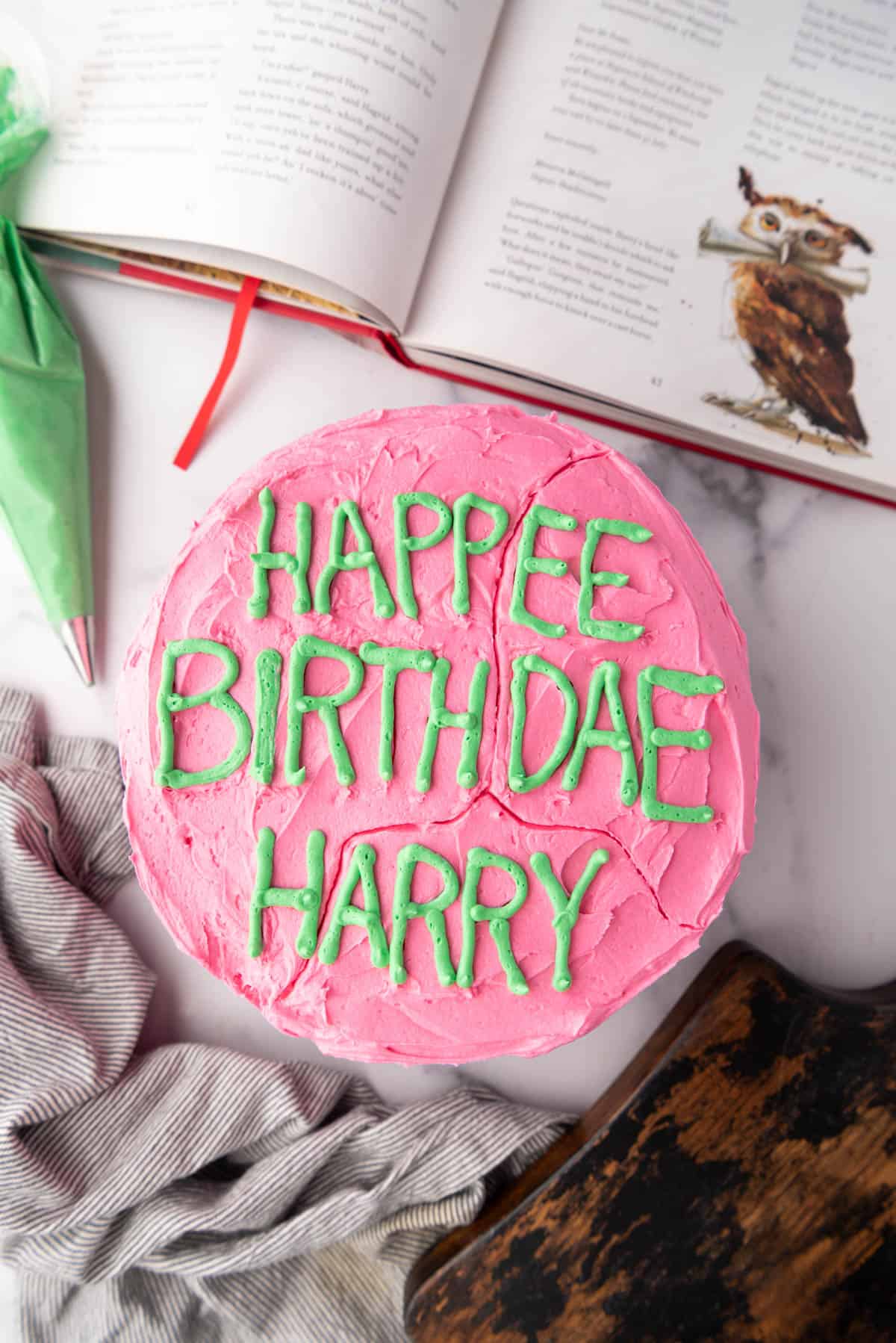 An overhead image of a Harry Potter birthday cake next to green frosting in a piping bag and an illustrated Harry Potter and the Philosopher's Stone book.