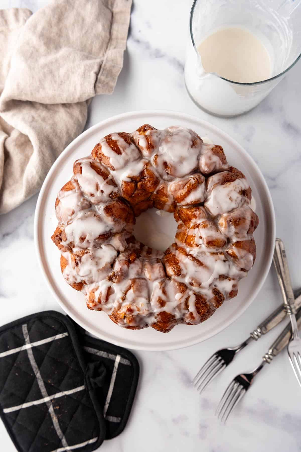 An overhead image of glazed cinnamon sugar monkey bread next to neutral linen napkins, a bowl of glaze, forks, and hot pads.