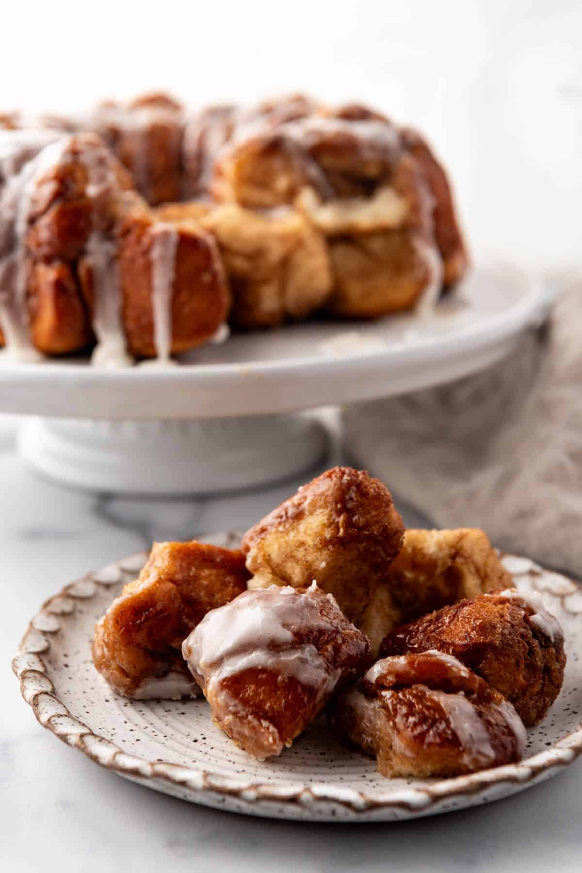 Pieces of sticky monkey bread on a plate in front of the rest of the monkey bread on a white cake stand.