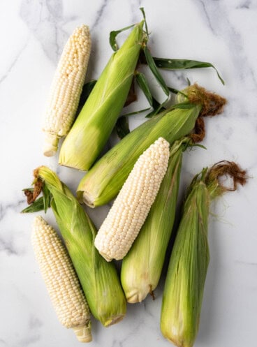 An overhead image of ears of corn on the cob with some of the in their green husks and others that have been shucked.