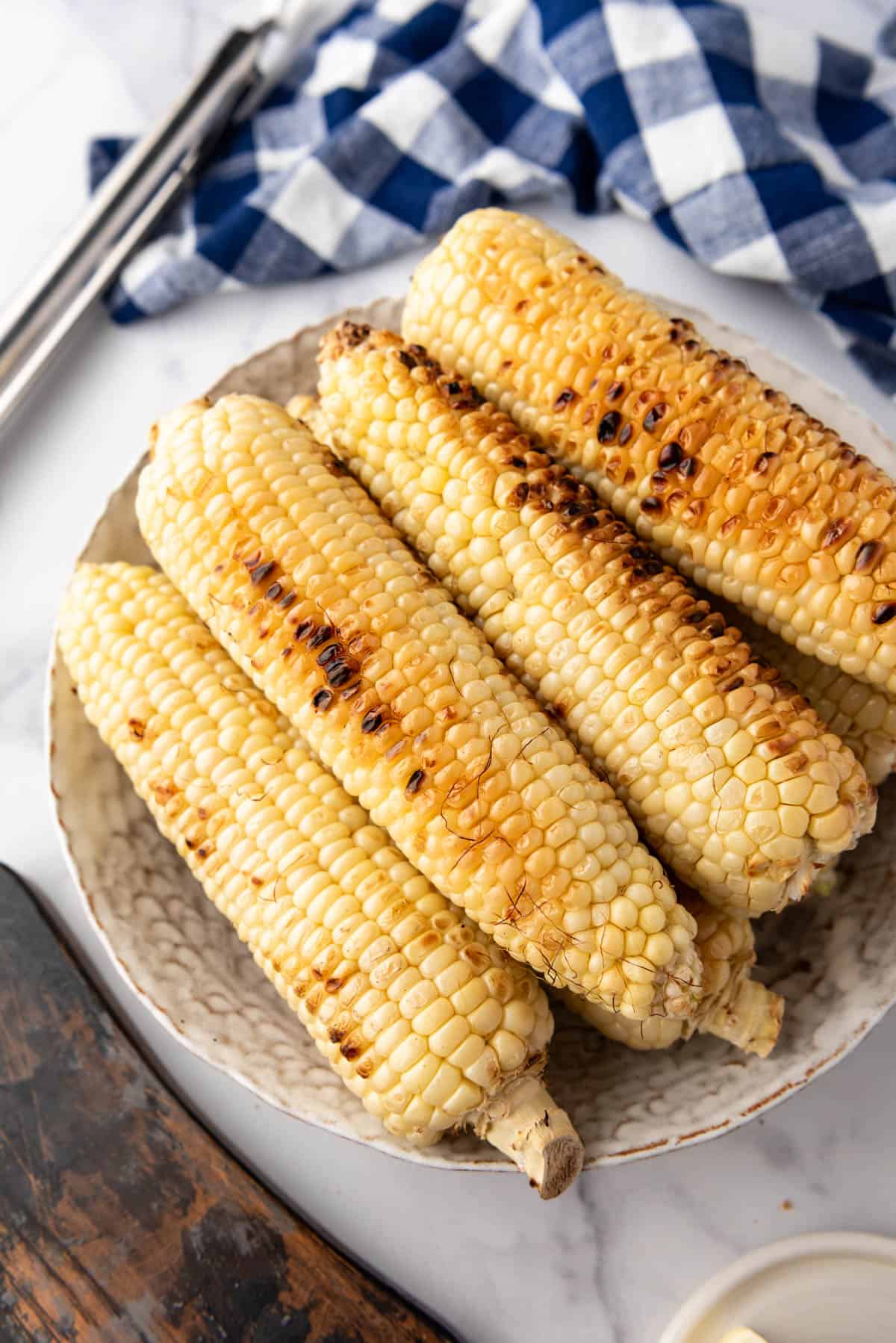 An overhead image of slightly charred ears of grilled corn on the cob with a blue and white checked cloth and tongs behind them.